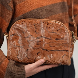 Op de afbeelding: Bruine schoudertas met slangenpatroon en lange riem. De tas wordt gedragen voor een persoon die een bruin-oranje gestreepte trui draagt.