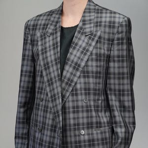 May include: A double-breasted blazer in a gray and black plaid pattern. The jacket has a classic design with notched lapels and multiple buttons. The fabric appears to be a woven material, suitable for a professional or vintage aesthetic.