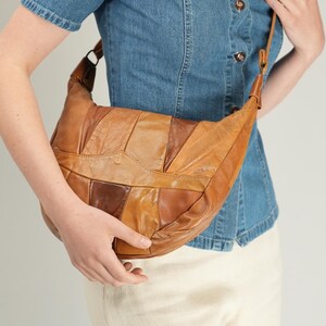 Pode incluir: Uma bolsa de ombro em forma de meia-lua construída com vários retalhos de couro em tons de castanho. A bolsa apresenta uma alça de couro longa e ajustável e uma silhueta curva, exibindo um design de patchwork com costuras visíveis entre as peças.