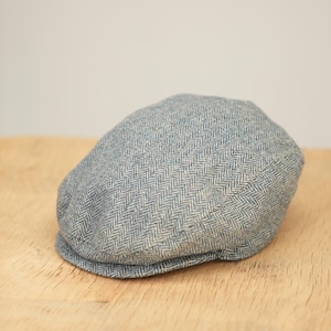 Puede incluir: Gorra plana azul claro con estampado de espiga. La gorra está hecha de una tela texturizada y tiene una corona redondeada y una pequeña visera rígida. La gorra está sobre una superficie de madera.
