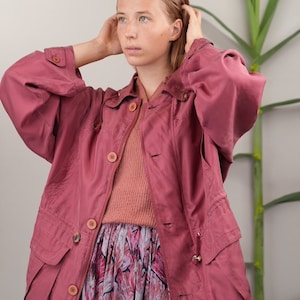 May include: A pink hooded jacket with a zipper and buttons. The jacket has two large pockets on the front and a drawstring at the waist. The model is wearing a pink sweater and a floral skirt underneath the jacket.