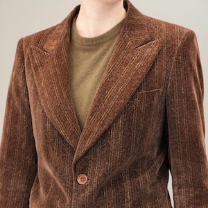 Puede incluir: Una chaqueta de pana marrón con solapa de muesca y tres botones. La chaqueta tiene un patrón a rayas y un bolsillo en el lado izquierdo.