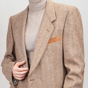 Könnte beinhalten: Ein brauner Tweed-Blazer mit Ellbogenpatches und Taschenakzenten aus Leder. Der Blazer hat ein klassisches Fischgrätenmuster und einen Zwei-Knopf-Verschluss. Die Person trägt einen beigen Rollkragenpullover und eine graue Hose.