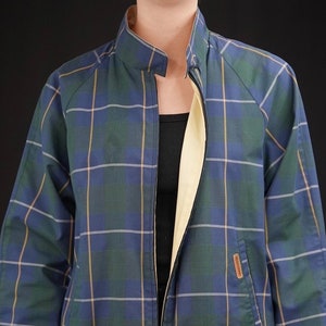 Può includere: Giacca a quadri blu, verdi e gialli con interno giallo chiaro. La giacca ha una chiusura con cerniera, un collo alto e polsini con bottoni. Una piccola tasca è visibile sul davanti.