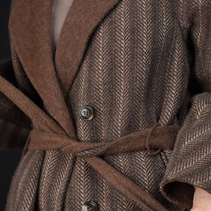 May include: A brown herringbone patterned coat with a brown self-tie belt. The coat has a double-breasted closure with two buttons.
