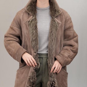 Könnte beinhalten: Ein langer brauner Wildledermantel mit einem Lammfellfutter. Der Mantel hat einen Knopfverschluss und zwei Taschen.