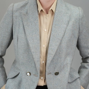 May include: A light blue blazer with a subtle herringbone pattern, featuring a notched lapel and two decorative buttons. The blazer is worn over a tan button-down shirt and black pants. A classic, tailored look.