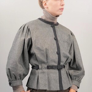 May include: A black and white houndstooth patterned jacket with a black belt and a dark gray trim. The jacket has a high collar and long sleeves.