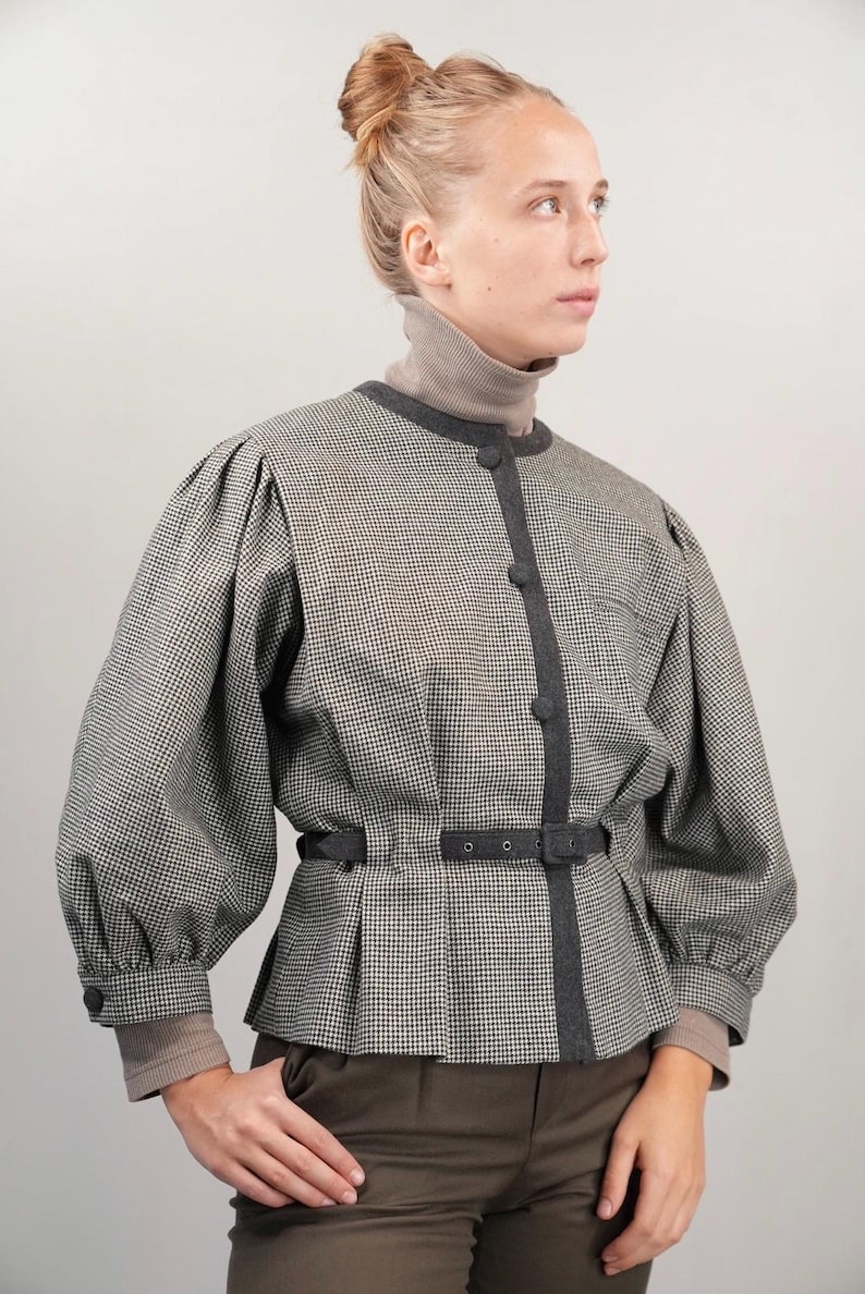 May include: A black and white houndstooth patterned jacket with a black belt and a dark gray trim. The jacket has a high collar and long sleeves.