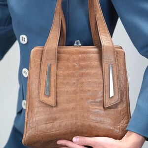 May include: A vintage brown leather handbag with a crocodile texture. The bag has two handles and silver-coloured metal accents. The bag is held against a blue background.