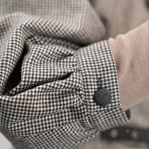 May include: A close-up of a black and white houndstooth patterned jacket with a black button. The jacket has a long sleeve and is made of a soft, textured fabric.