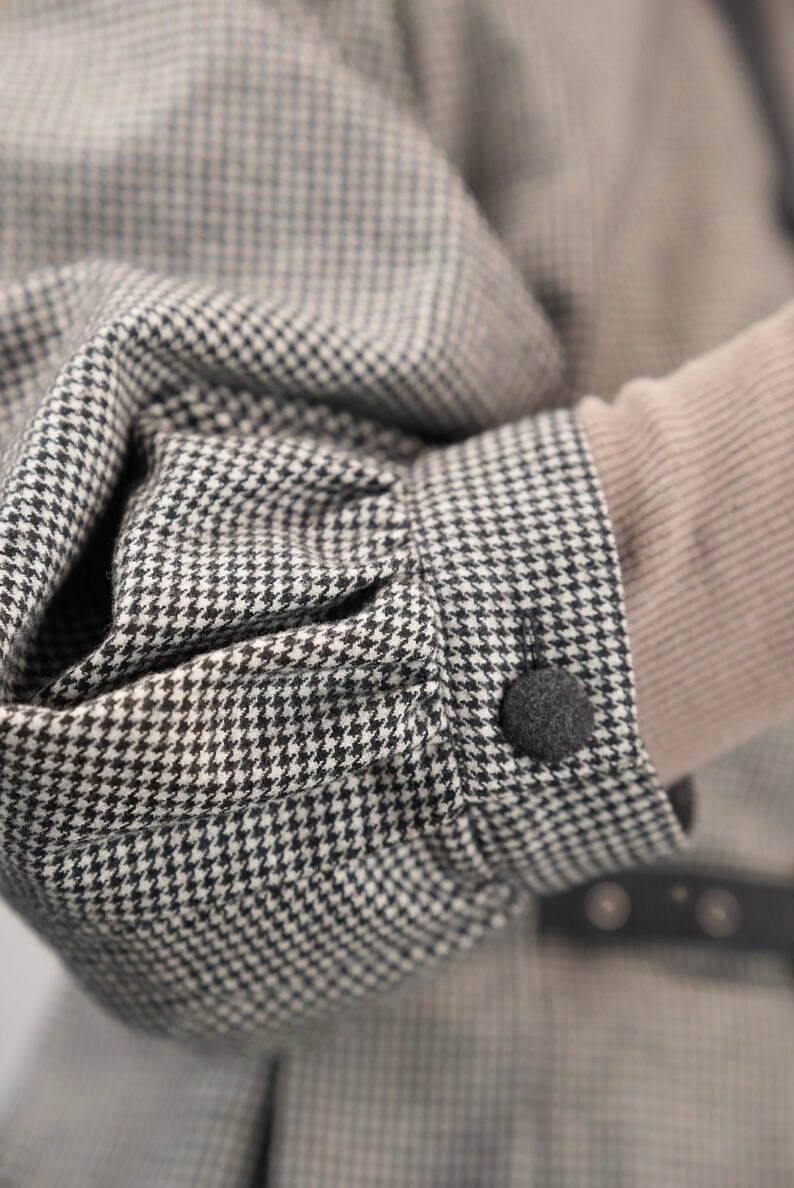 May include: A close-up of a black and white houndstooth patterned jacket with a black button. The jacket has a long sleeve and is made of a soft, textured fabric.