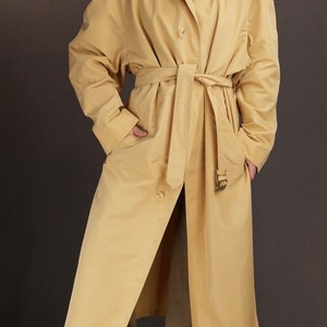 May include: A long, yellow trench coat with a belt and pockets. The coat is made of a smooth, lightweight fabric.