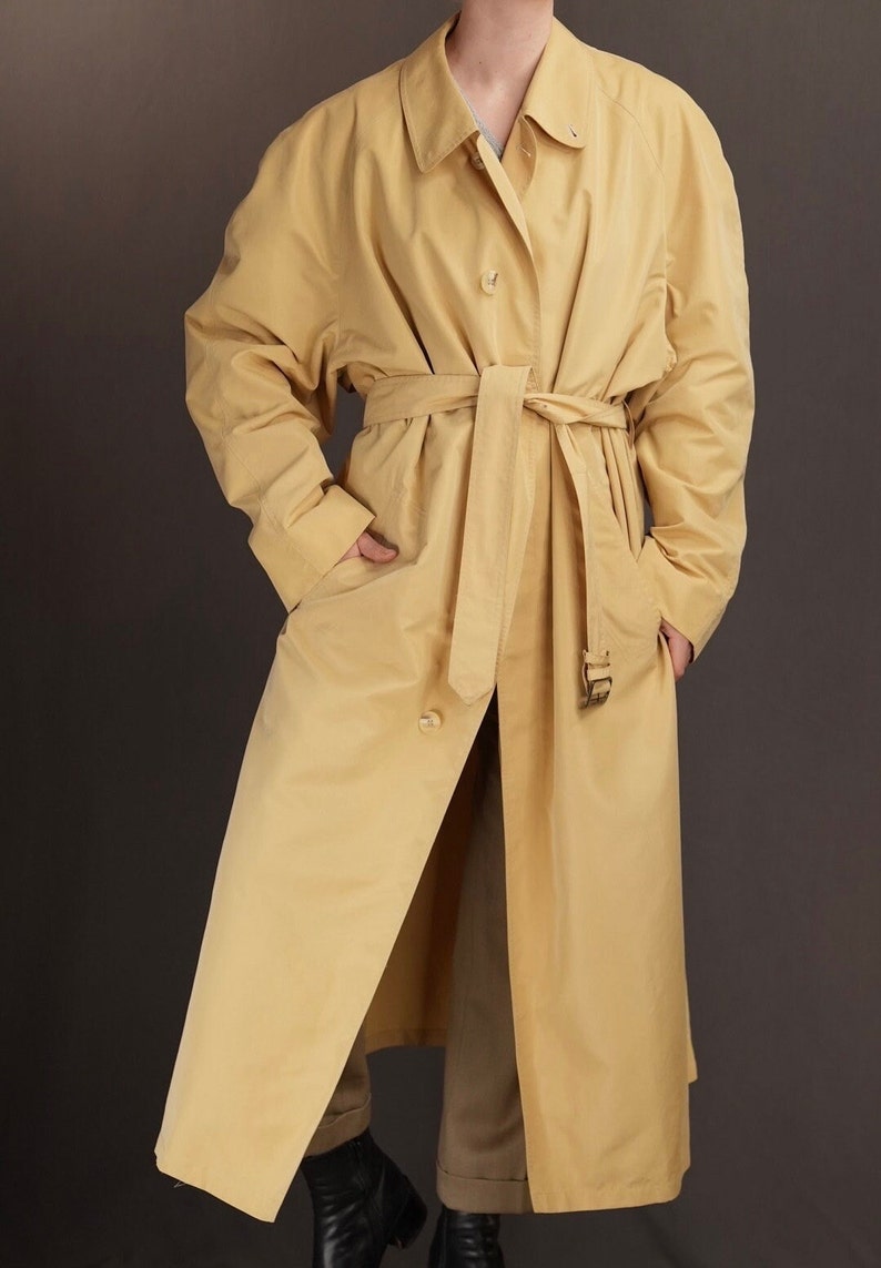 May include: A long, yellow trench coat with a belt and pockets. The coat is made of a smooth, lightweight fabric.
