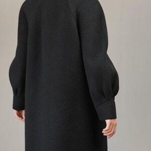 May include: A black long-sleeved coat with a high mock neck collar and a slightly oversized fit. The coat has a textured fabric and a slightly gathered detail at the shoulders.