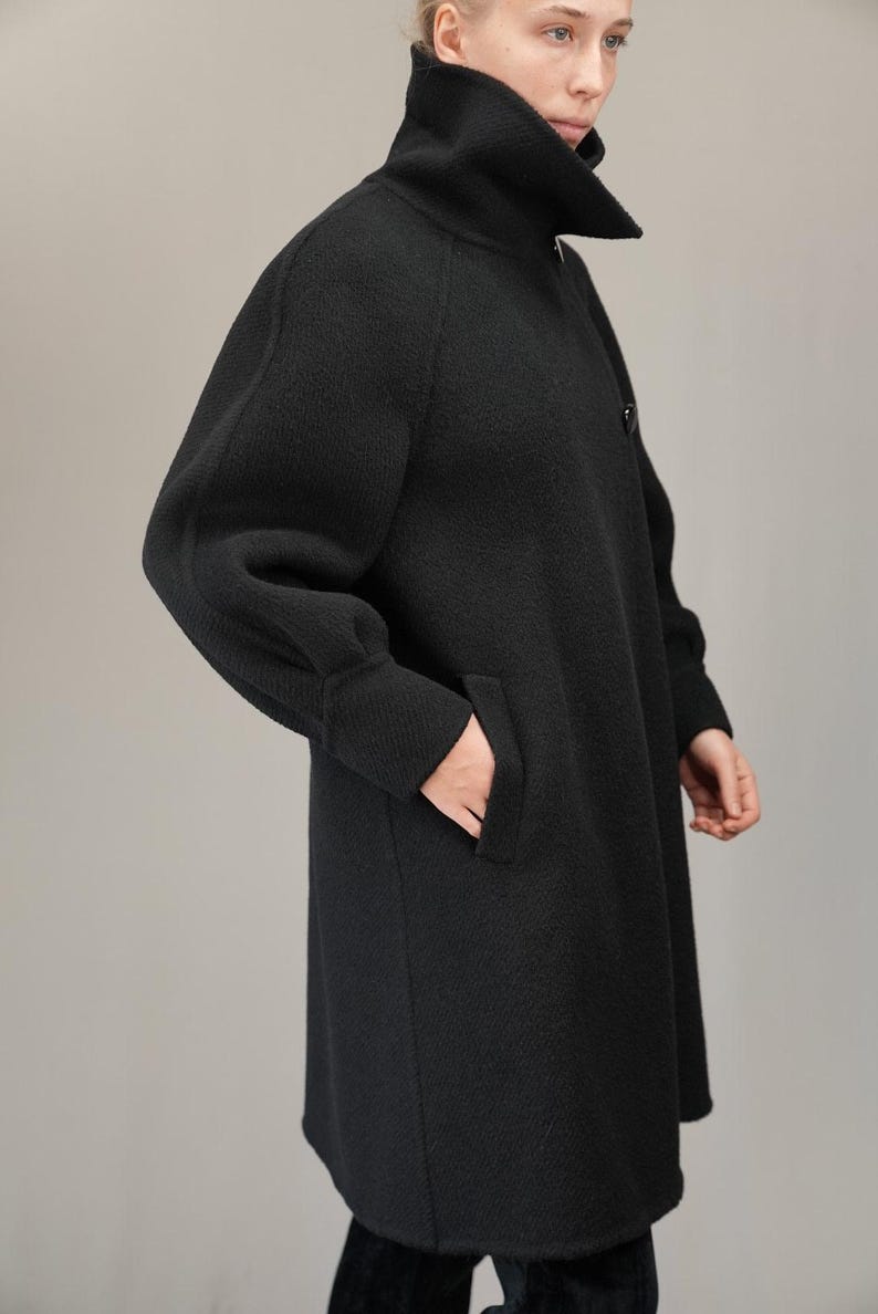 May include: A black long-sleeved coat with a high collar and a single button closure. The coat has a relaxed fit and a slightly flared silhouette.