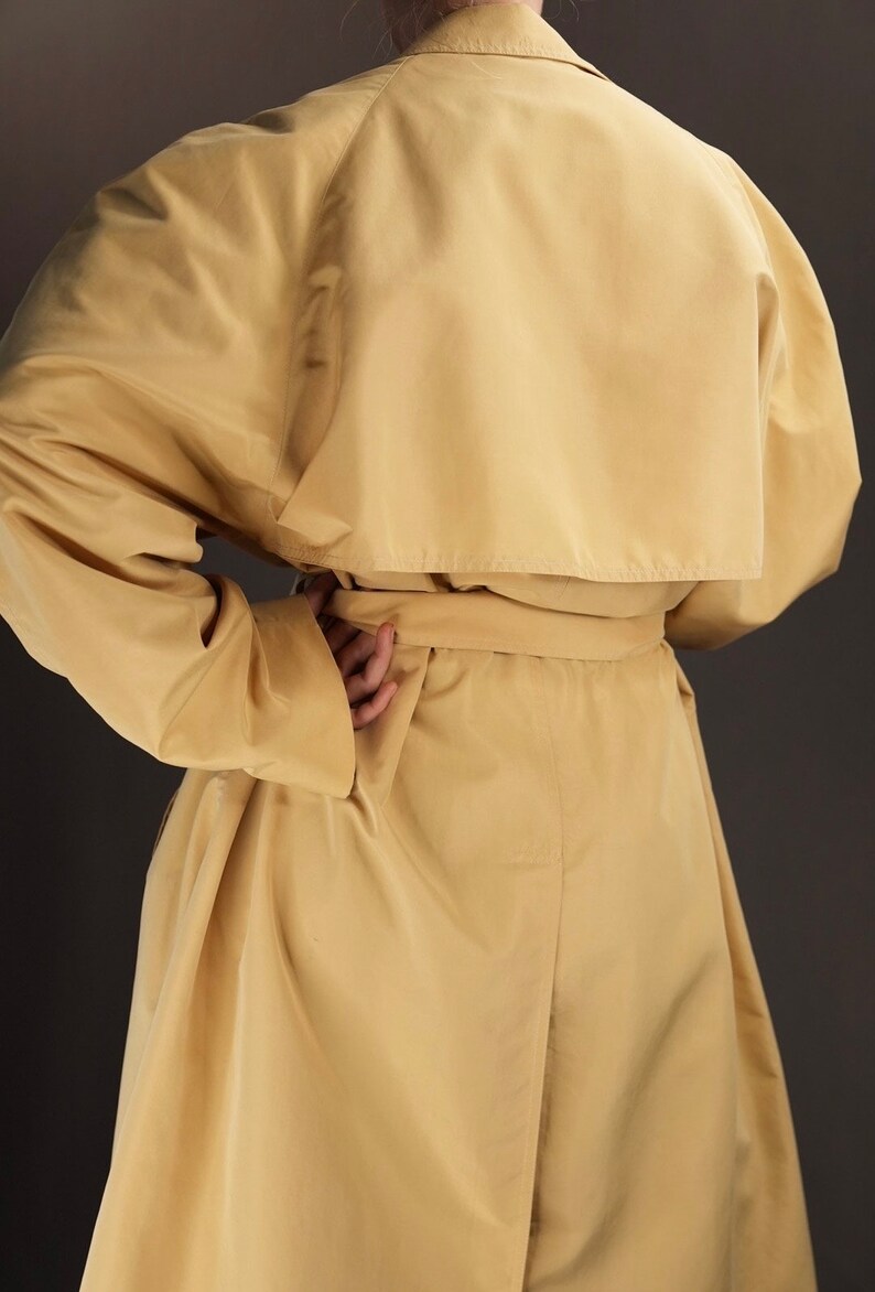 May include: A long, light yellow trench coat with a belt. The coat has a classic design with a collar and lapels.