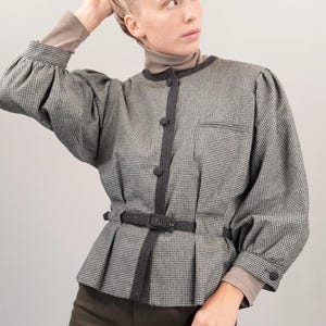 May include: A black and white houndstooth patterned blouse with a black belt and gray trim. The blouse has a button closure and long sleeves.