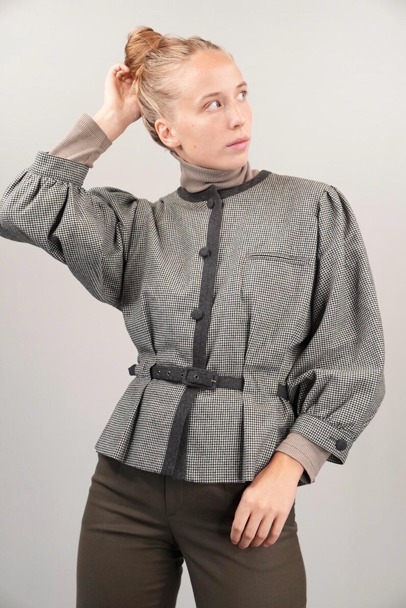 May include: A black and white houndstooth patterned blouse with a black belt and gray trim. The blouse has a button closure and long sleeves.