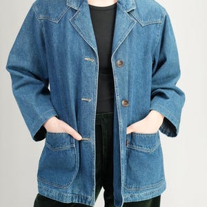May include: A medium-wash denim jacket with a classic collar, button closure, and two front pockets. The jacket has a relaxed fit and is worn over a black top and dark green pants. The jacket's buttons are a brown color.