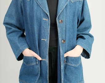 May include: A medium-wash denim jacket with a classic collar, button closure, and two front pockets. The jacket has a relaxed fit and is worn over a black top and dark green pants. The jacket's buttons are a brown color.
