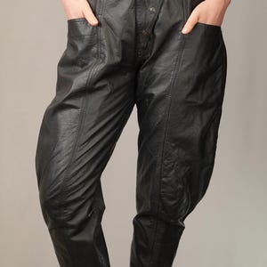 May include: Black leather pants with a high waist and a button fly. The pants have a unique design with seams running down the legs and a cinched ankle.