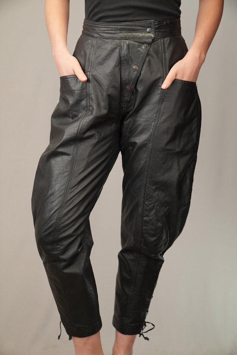 May include: Black leather pants with a high waist and a button fly. The pants have a unique design with seams running down the legs and a cinched ankle.