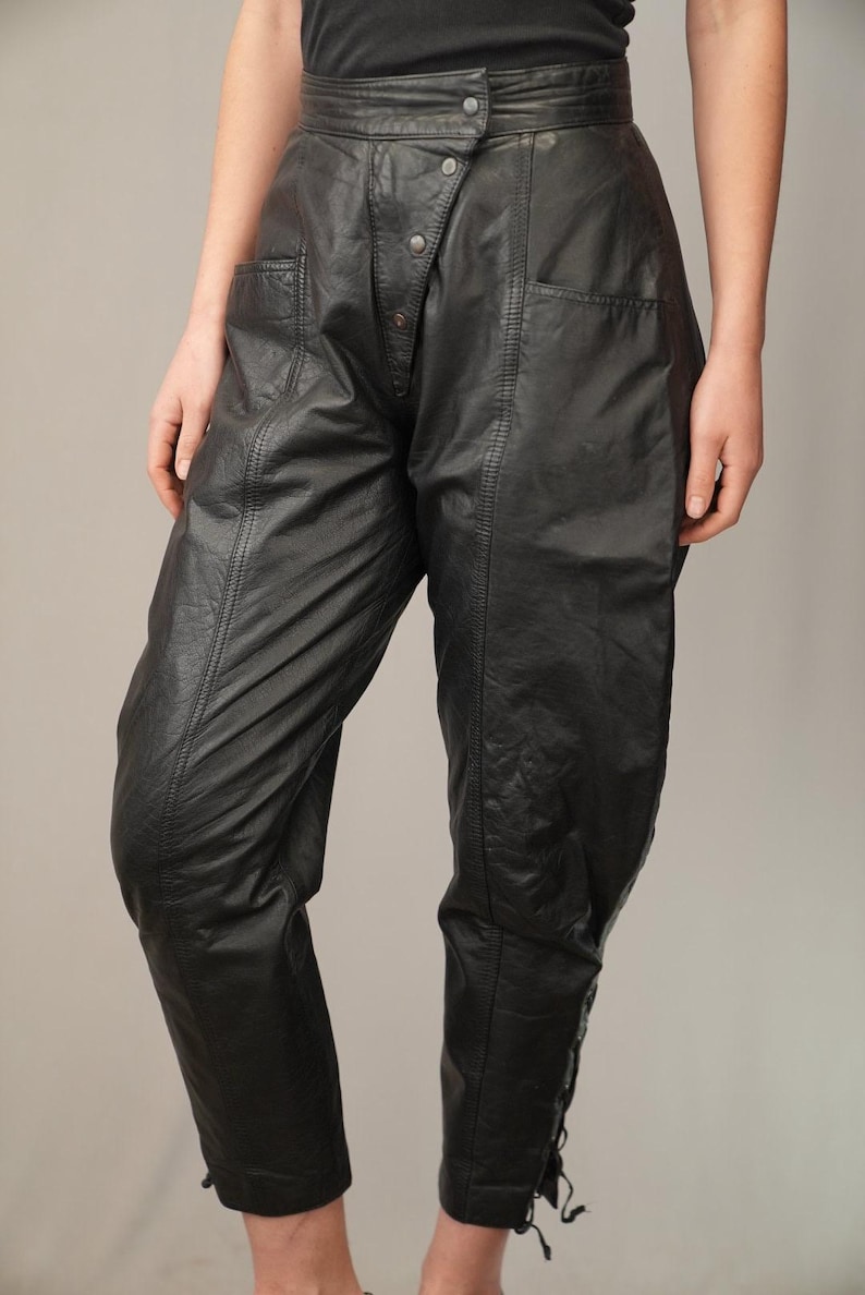 May include: Black leather pants with a high waist and a button fly. The pants have a unique design with a diagonal seam and a flap pocket on each side.