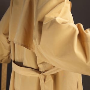 May include: A beige trench coat with a belt tied at the waist. The coat has a collar and a single breasted closure.