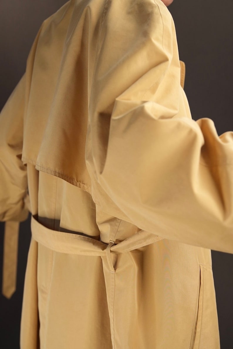 May include: A beige trench coat with a belt tied at the waist. The coat has a collar and a single breasted closure.