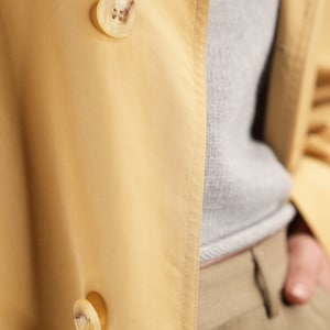 May include: A yellow trench coat with two large, light brown buttons. The coat is made of a smooth, silky fabric.