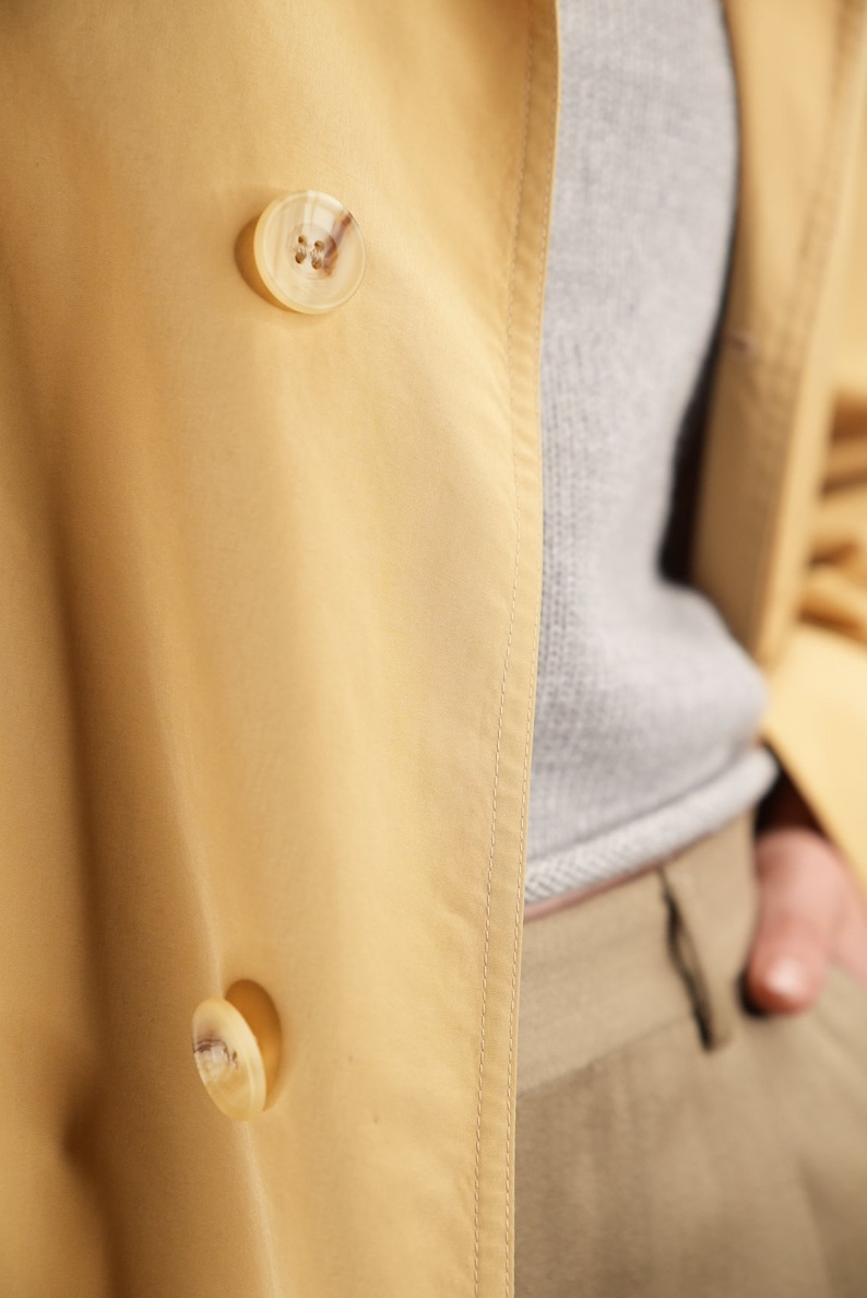 May include: A yellow trench coat with two large, light brown buttons. The coat is made of a smooth, silky fabric.