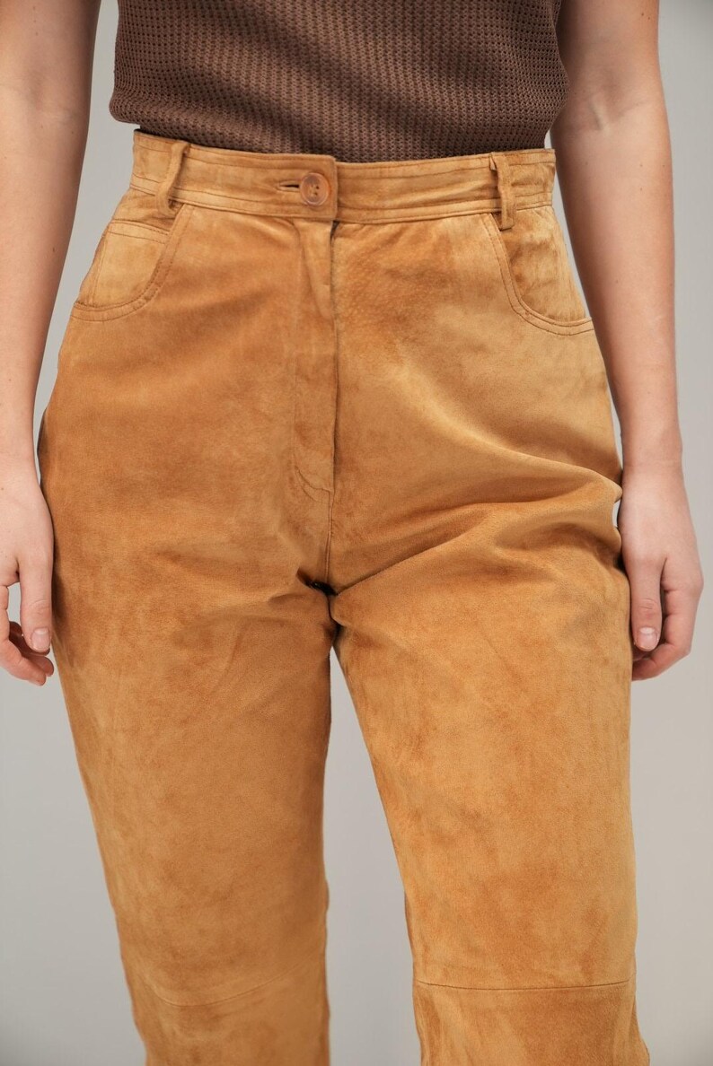May include: A pair of high-waisted brown suede pants with a button and zipper closure. The pants have a relaxed fit and are made of a soft, supple suede.