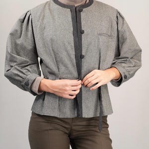 May include: A black and white houndstooth patterned jacket with a dark gray trim and a tie closure. The jacket has long sleeves and a round neckline.