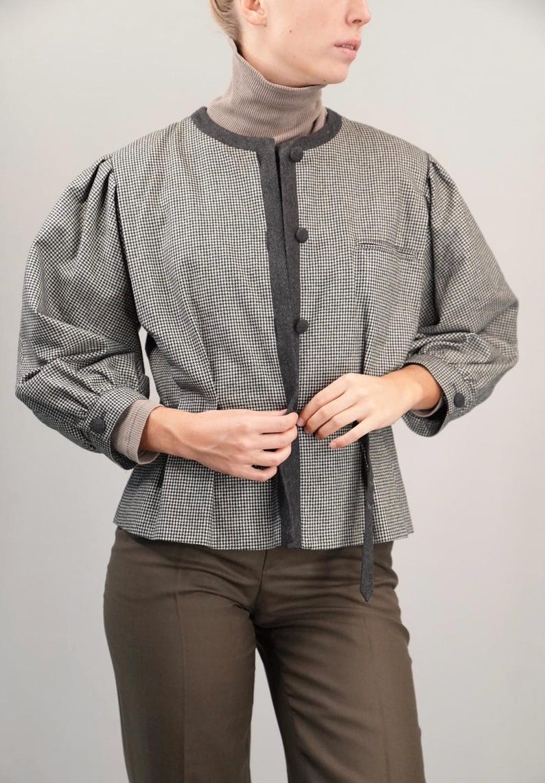 May include: A black and white houndstooth patterned jacket with a dark gray trim and a tie closure. The jacket has long sleeves and a round neckline.