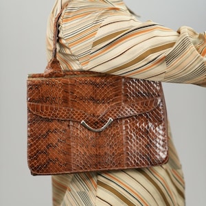 Pode incluir: Bolsa de mão retangular marrom com padrão de pele de cobra e fecho prateado. A bolsa tem uma alça e uma aba. O fundo inclui uma camisa listrada em tons de marrom, laranja e creme.