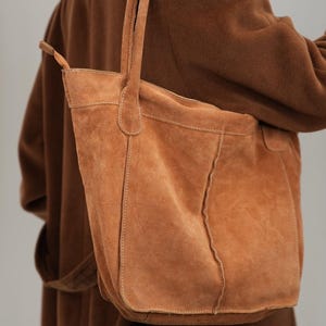 Könnte beinhalten: Eine hellbraune Wildleder-Tragetasche mit rechteckiger Form und zwei Griffen. Die Tasche hat sichtbare Nähte und eine weiche Textur. Die Tasche wird über der Schulter getragen, und die Person trägt einen braunen Mantel.