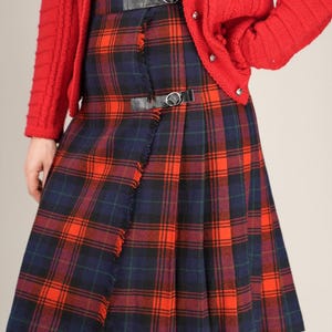 May include: A red cardigan worn over a tartan skirt with a black belt. The skirt displays a classic plaid pattern in red, navy, and green. The skirt has a fringed edge and is pleated.