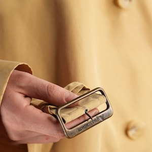 May include: A close-up of a beige trench coat with a brass buckle. The buckle has the text "Andreas Murr" engraved on it.