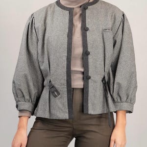 May include: A black and white houndstooth patterned jacket with a dark gray trim and a brown turtleneck sweater underneath. The jacket has a cinched waist and long sleeves.