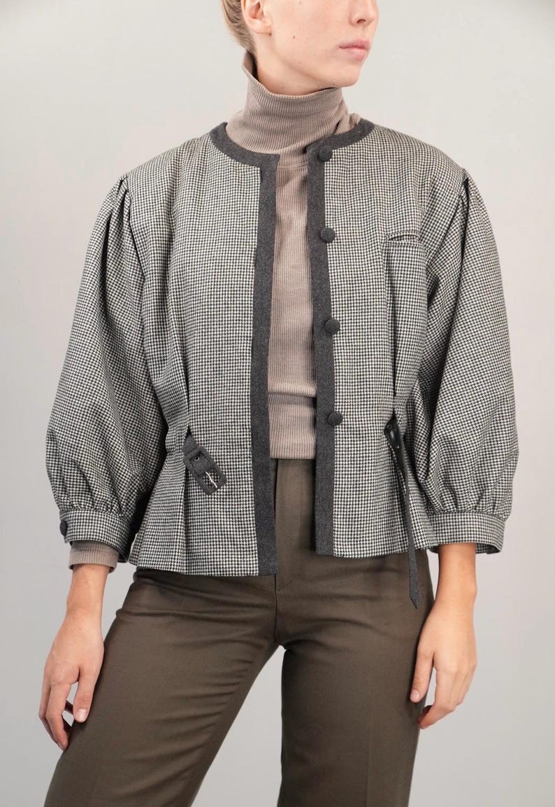 May include: A black and white houndstooth patterned jacket with a dark gray trim and a brown turtleneck sweater underneath. The jacket has a cinched waist and long sleeves.