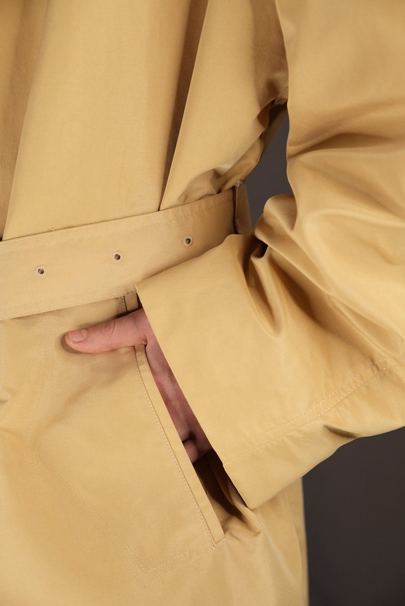 May include: A beige trench coat with a belt and a pocket. The coat is made of a smooth, lightweight fabric.
