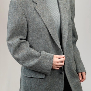 May include: A grey tweed blazer with a notched lapel, two front pockets, and a single-button closure. The blazer is worn over a light grey ribbed turtleneck. The fabric has flecks of brown and other colours.