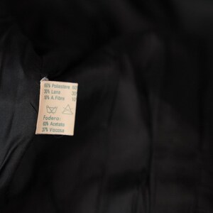 May include: A white fabric label with black text detailing the fabric composition of a garment. The label reads: "60% Poliestere 60, 20% Lana, 10% A Fibre 10, Fodera, 60% Acetato, 37% Viscosa".