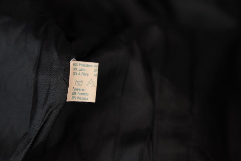 May include: A white fabric label with black text detailing the fabric composition of a garment. The label reads: "60% Poliestere 60, 20% Lana, 10% A Fibre 10, Fodera, 60% Acetato, 37% Viscosa".