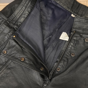 May include: Black leather pants with a zipper and multiple buttons. The pants have a flap pocket on the right side.