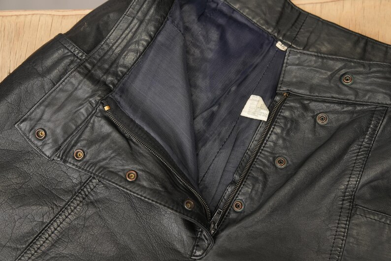 May include: Black leather pants with a zipper and multiple buttons. The pants have a flap pocket on the right side.