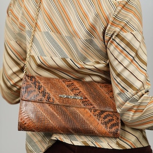 Op de afbeelding: Een vintage clutch met een slangenleerpatroon in bruin- en oranjetinten. De tas heeft een zilverkleurige sluiting en een gouden kettingriem. De achtergrond toont een gestreepte blouse in beige, bruin en oranje.
