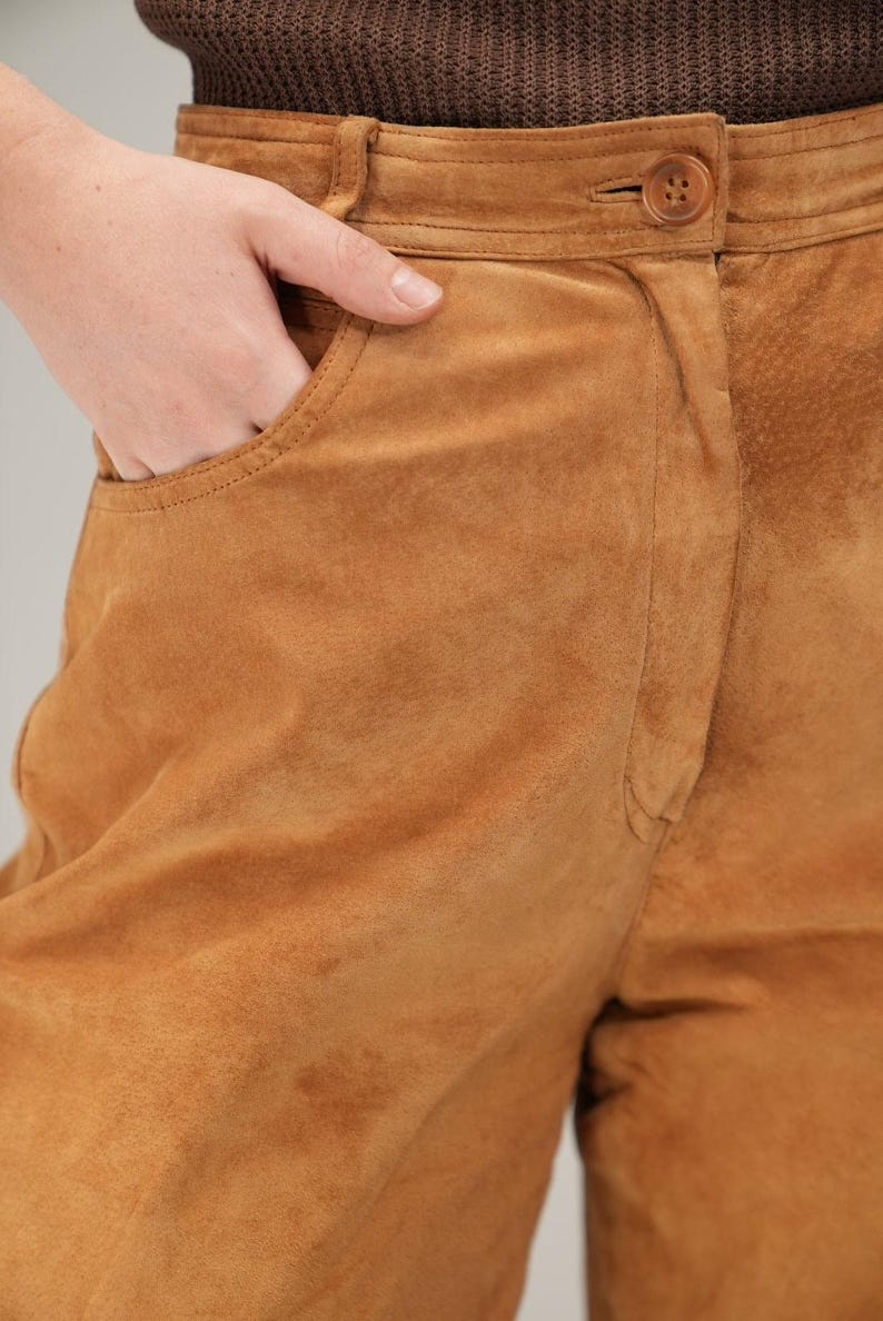 May include: Brown suede pants with a high waist and a button closure. The pants have a front pocket and a relaxed fit.