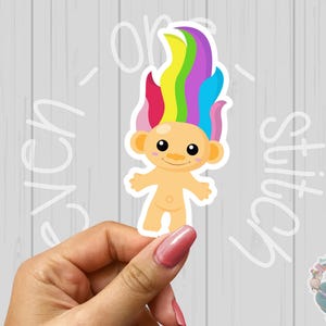 May include: A sticker of a cartoon troll with rainbow hair. The troll is smiling and has a light brown skin tone. The sticker is on a white background.